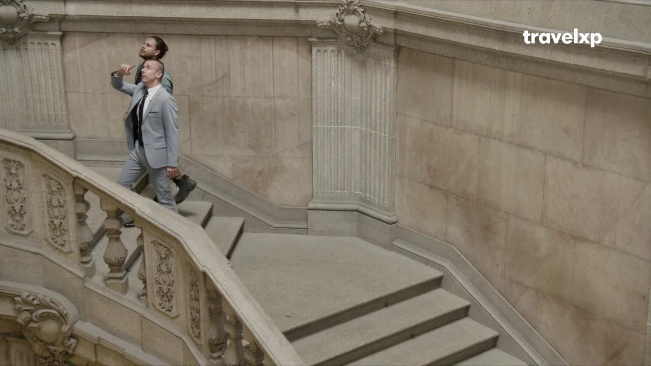 Two men in matching gray suits are ascending a grand, stone staircase. The man in front carries a briefcase, while the other points upwards, as if directing attention.

