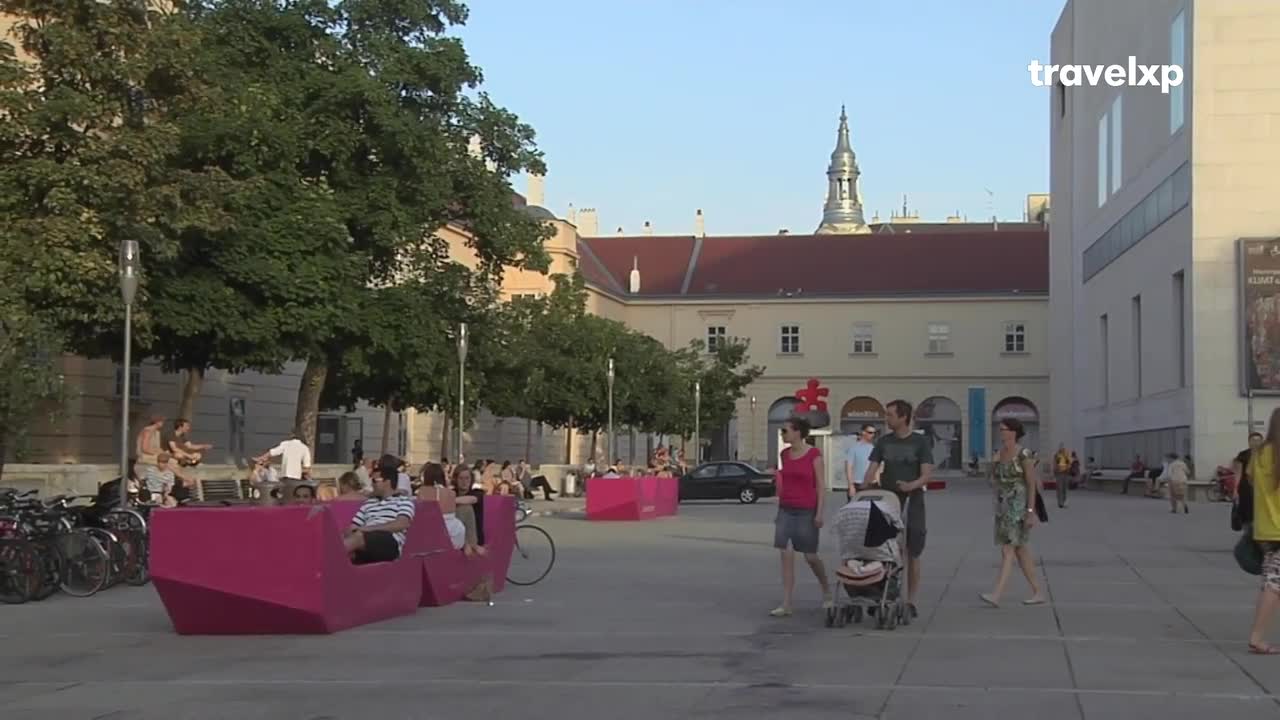 People stroll across a wide, paved plaza, some pushing strollers or sitting on bright pink benches. In the background, a couple of musicians play guitar for a small crowd gathered near the trees.
