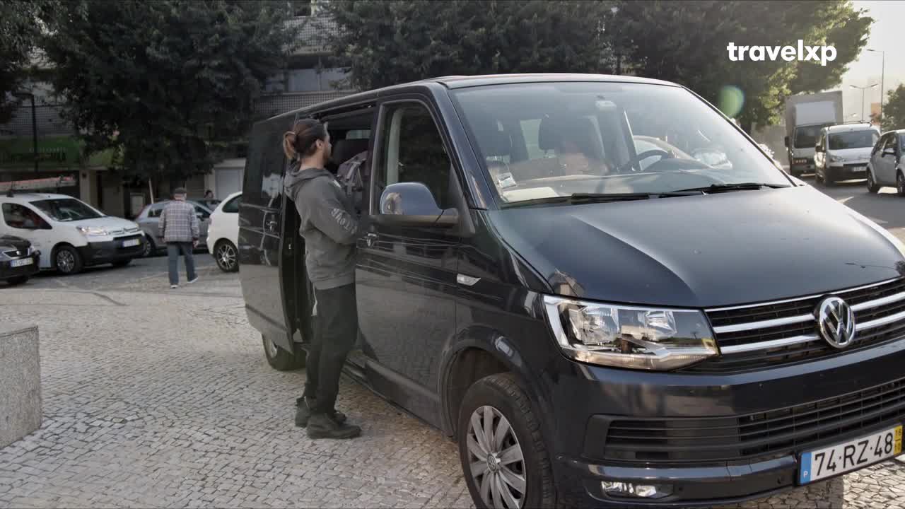 A man in a gray hoodie stands by the open side door of a dark van, seemingly waiting.  The van is parked on a cobblestone street, with other vehicles and buildings in the background.
