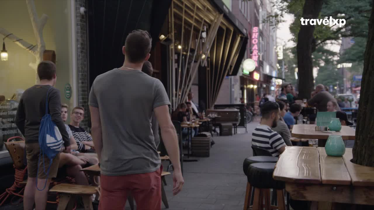 People are milling around a busy outdoor cafe, some seated at tables, others walking along the sidewalk. The neon sign of a "SHOARMA" restaurant glows in the fading light.
