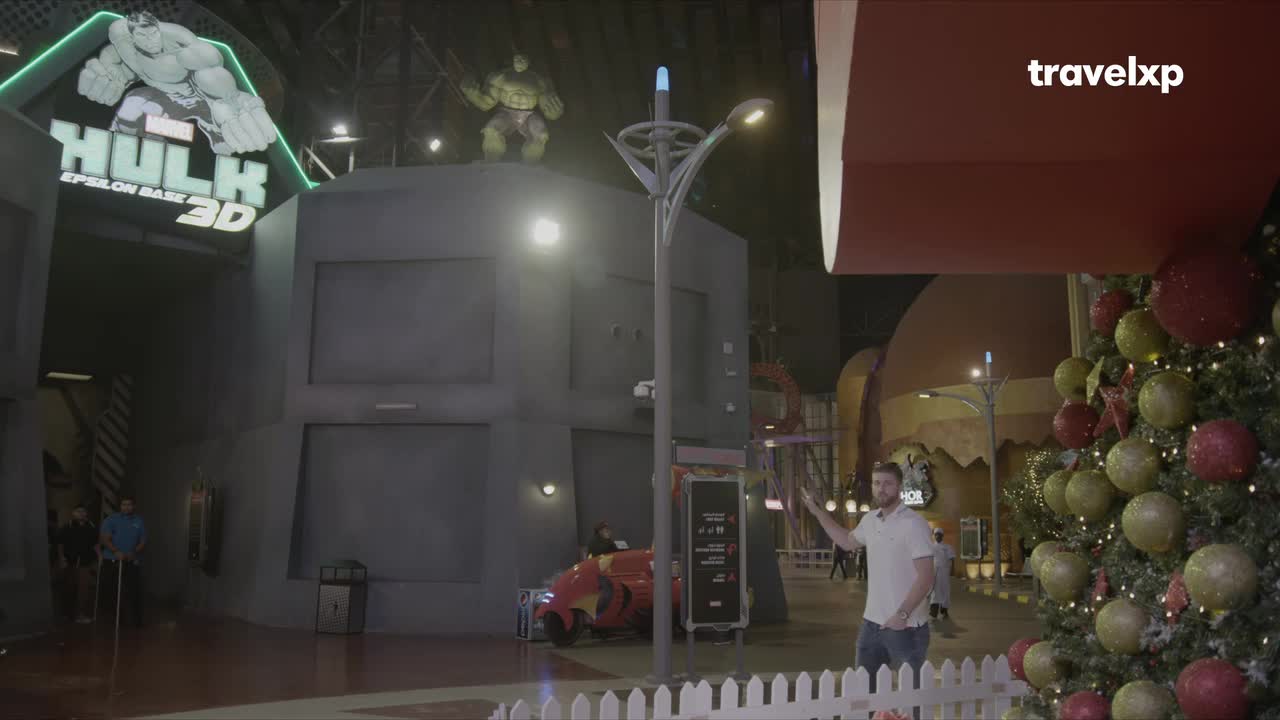 A man gestures toward a Hulk-themed attraction, with a giant figure of the hero perched above the entrance. Festive decorations, including a Christmas tree, add a surprising touch to the scene, possibly from India, as seen on Travelxp.
