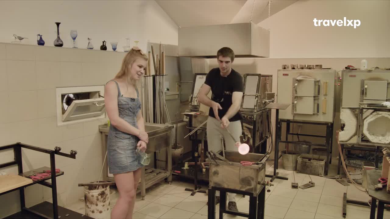 A glassblower, holding a long metal rod, is shaping a glowing, molten bubble. A woman in a denim dress watches, smiling, as the Travelxp show films the process.
