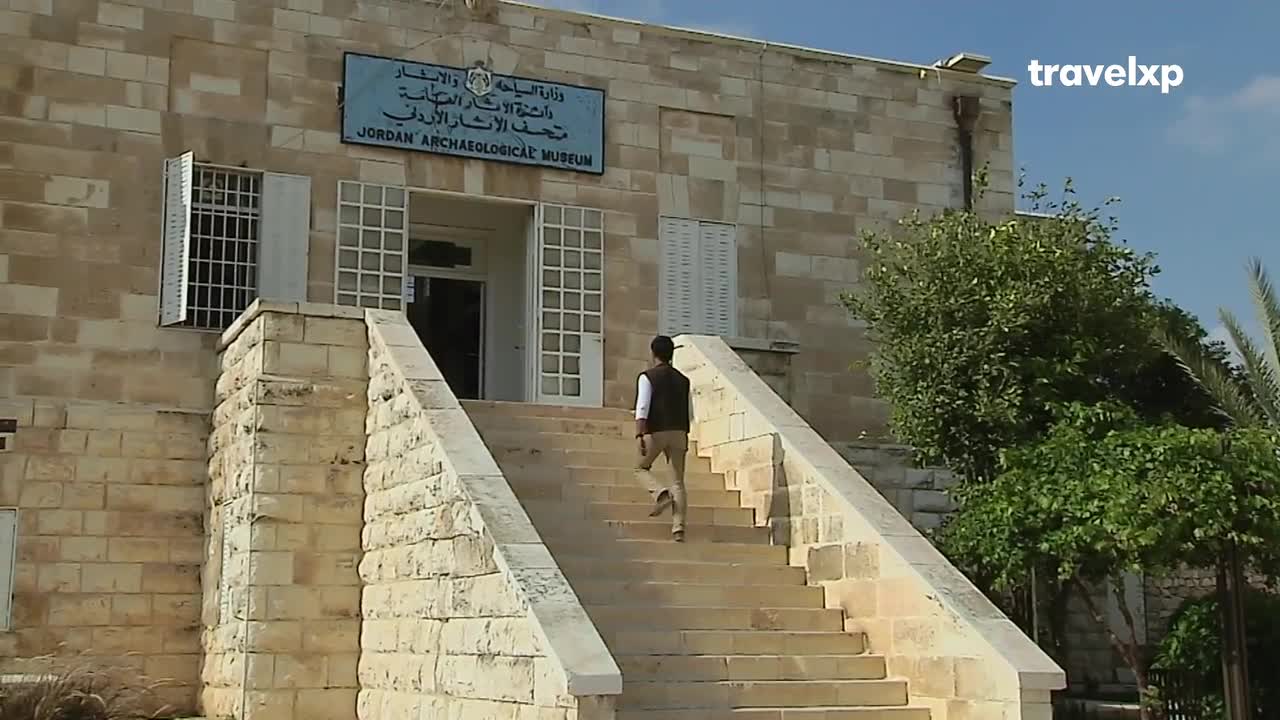 A man in a vest and trousers strides up the stone steps toward the Jordan Archaeological Museum. The building's sign, with Arabic script, sits above the open doorway.
