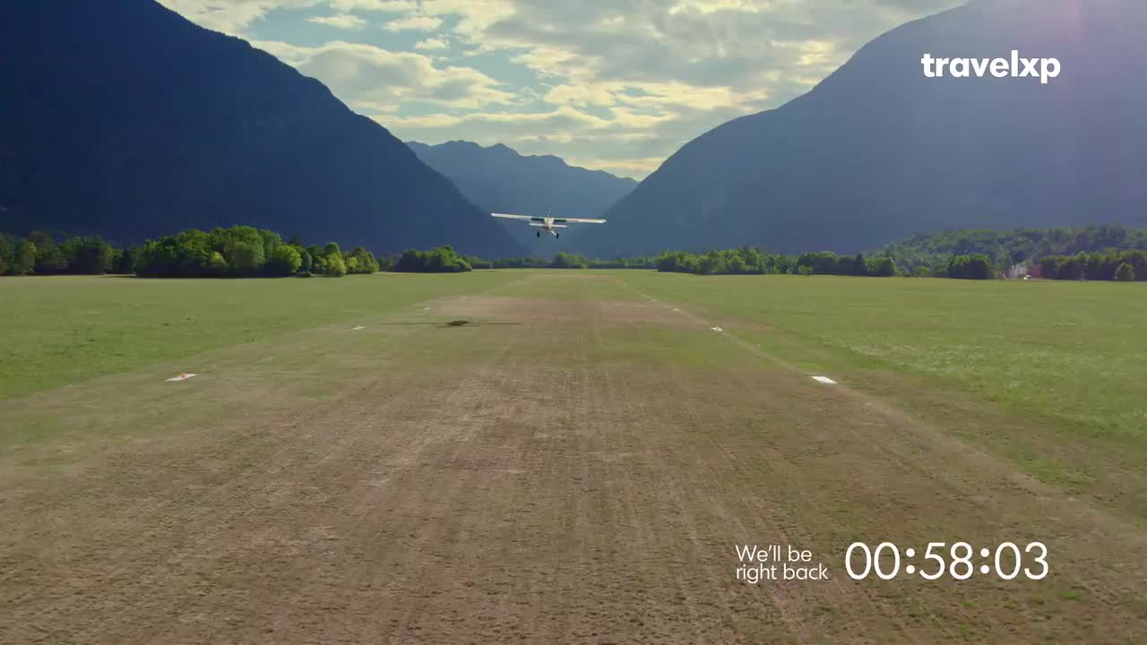 A small plane is descending towards a grassy airstrip, framed by towering mountains. The Travelxp logo and a countdown timer appear, signaling a break in the program.
