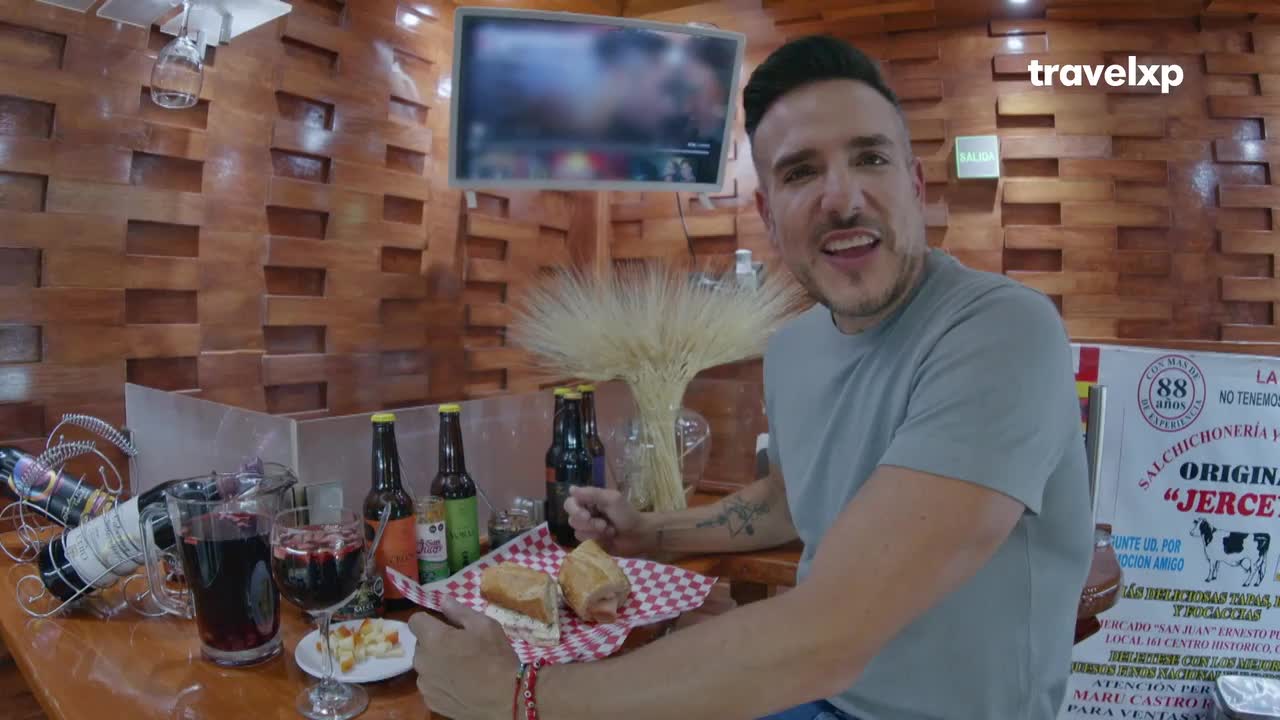 A man grins at the camera, gesturing toward a sandwich and a glass of red wine. Behind him, a wall of wooden planks and a Travelxp logo suggest a food-focused travel show.

