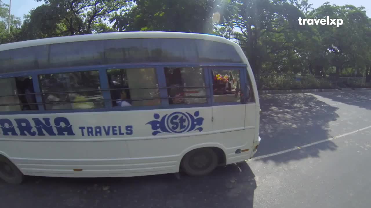 A white bus, marked "Karna Travels," rolls slowly on the asphalt. The Travelxp logo sits in the top right corner, and the bus windows reflect the bright sunlight.

