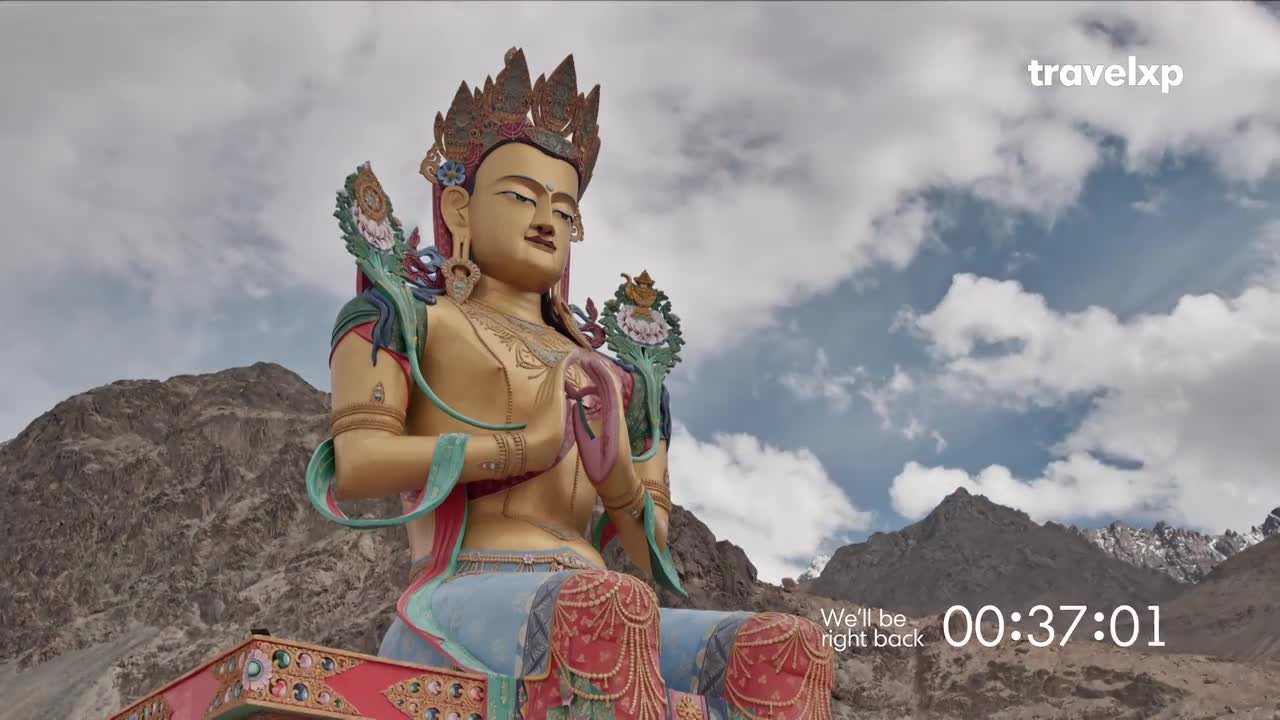 A massive, golden statue of a seated Buddha dominates the frame, set against a backdrop of rugged mountains and a cloudy sky. The Travelxp logo and a countdown timer appear, indicating a break in programming.
