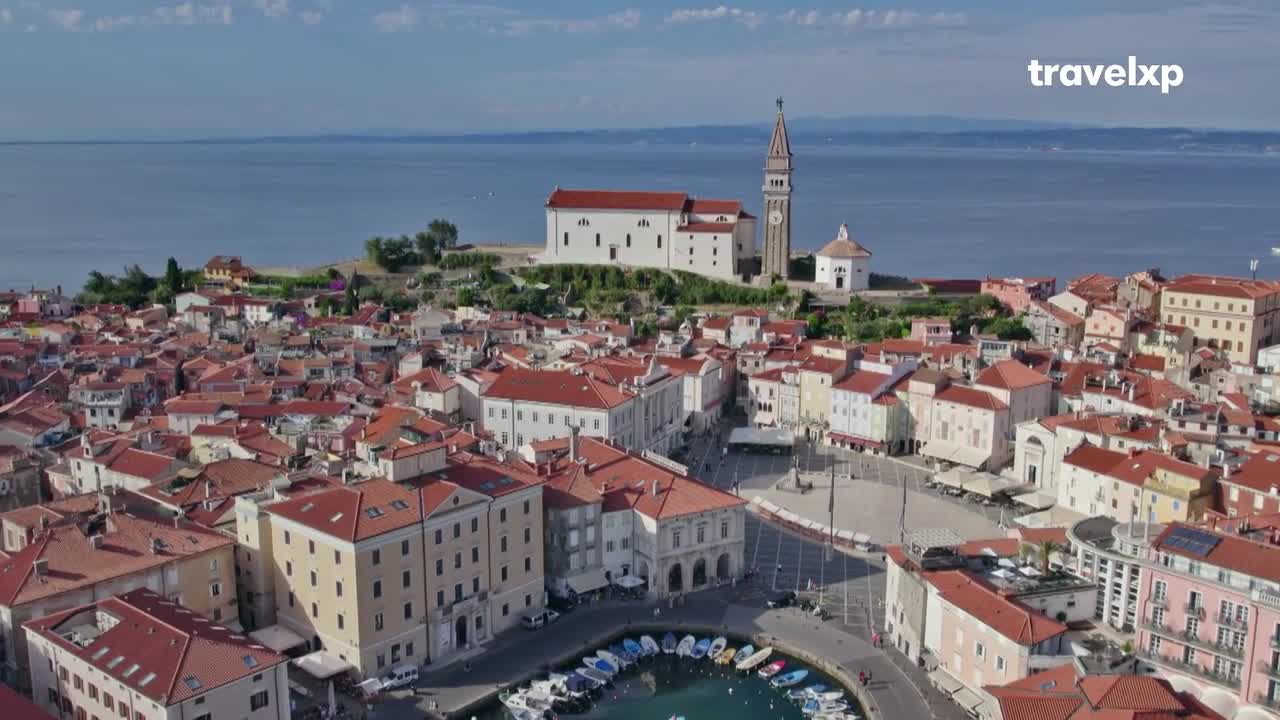 From this height, the terracotta rooftops of the town stretch toward the sea, punctuated by a tall church tower. The water is calm, and a few boats bob in the harbor below.
