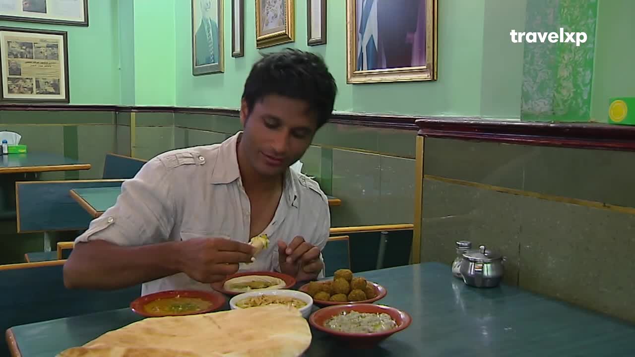 A man in a light shirt is holding a small piece of food, about to eat it from a plate of various dishes. He's sitting at a table in a restaurant, with a large piece of flatbread in the foreground.
