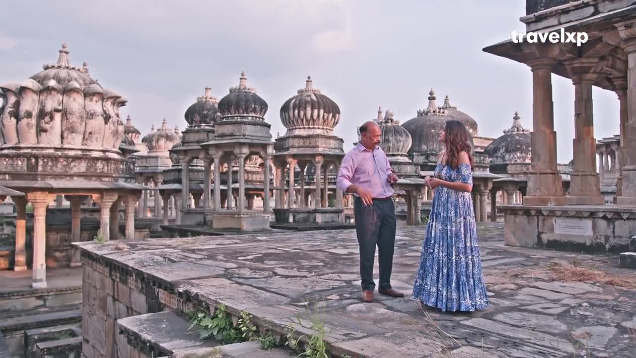 A man in a pink shirt gestures as he speaks to a woman in a blue dress, both standing on a stone platform. Behind them, a series of domed structures stretch across the horizon in India, likely for a Travelxp show.

