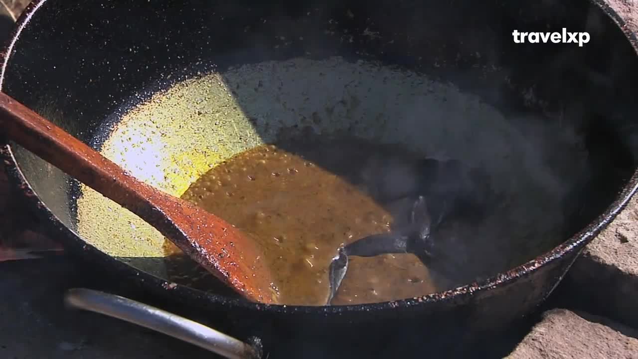 A wooden spoon stirs a thick, bubbling mixture in a large, blackened pot. Steam rises from the cooking food, illuminated by a patch of sunlight.
