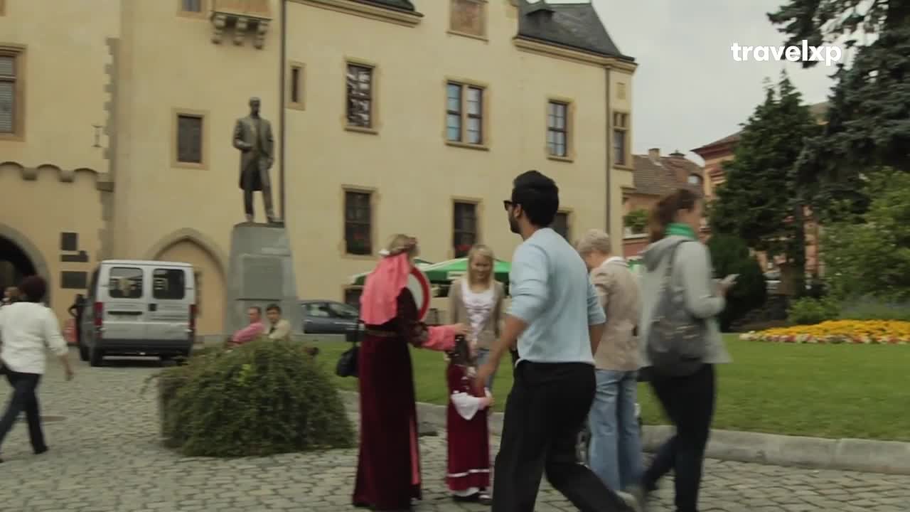 A man in sunglasses walks past a woman in a red medieval costume, who is handing something to another woman. Behind them, a group of people stroll on the cobblestone path near a large building.
