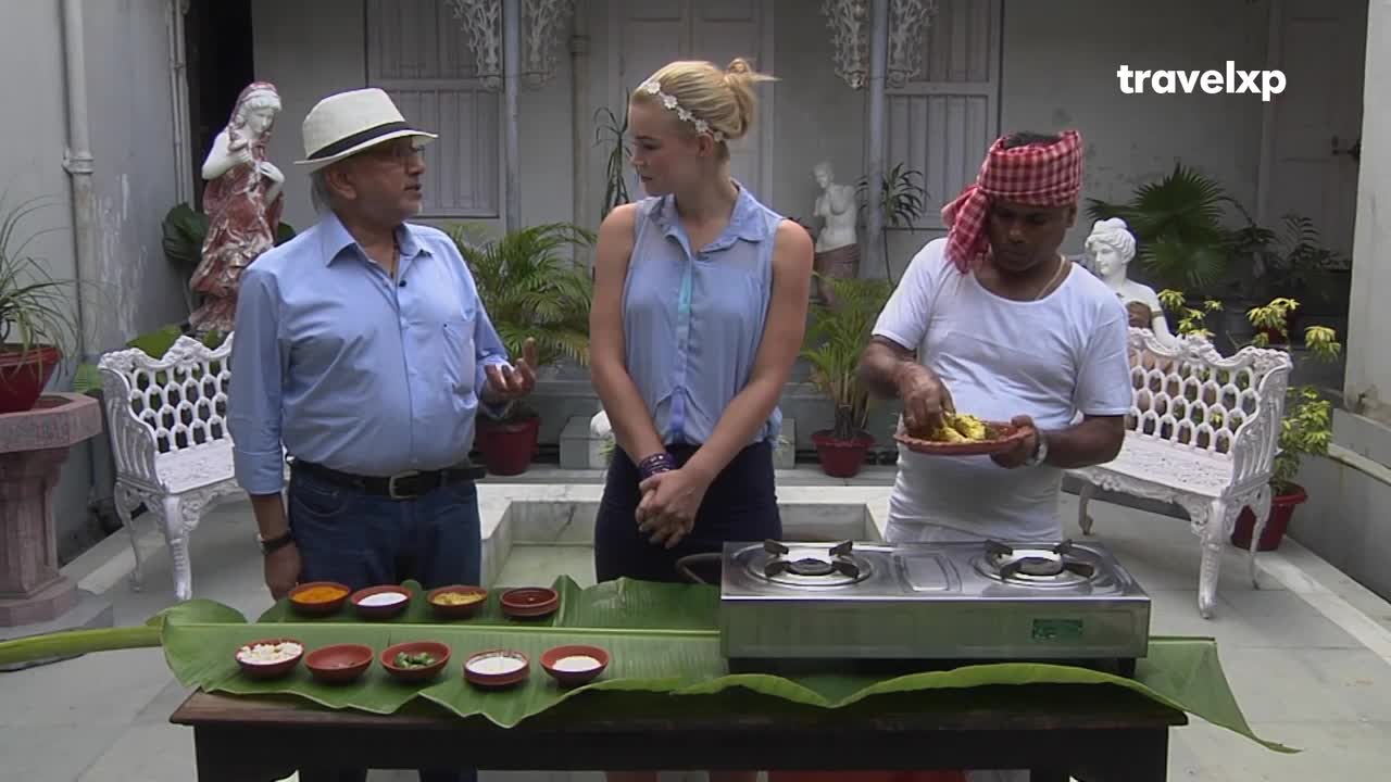 A man in a white hat gestures, explaining something to a woman as a cook in a red and white headscarf prepares food. A table is set with small bowls of spices and ingredients, all arranged on a large banana leaf.
