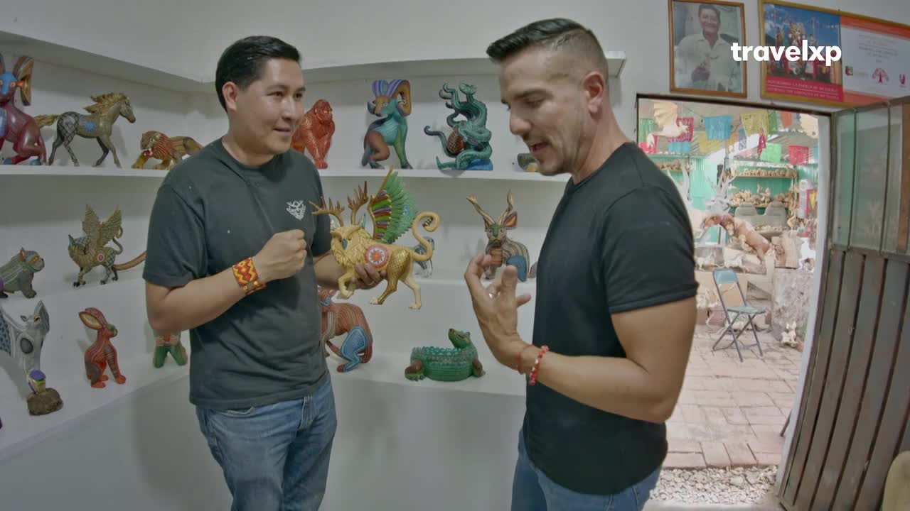 Two men stand in a workshop, gesturing towards colorful, fantastical wooden sculptures. The man on the right holds a small creature, while the other displays a winged deer, both surrounded by similar creations on shelves.
