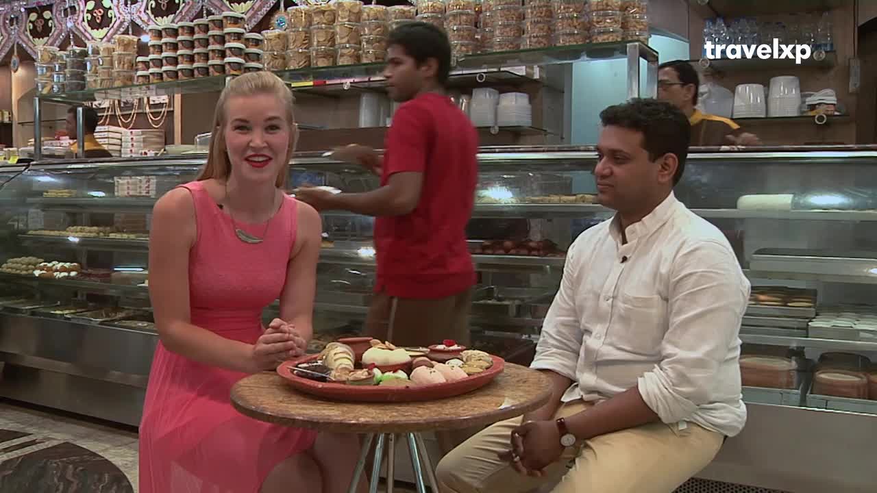 A woman in a pink dress smiles at the camera, while a man in a white shirt sits beside her. Behind them, a server in a red shirt is arranging items in a glass display case, possibly for a Travelxp segment in India.
