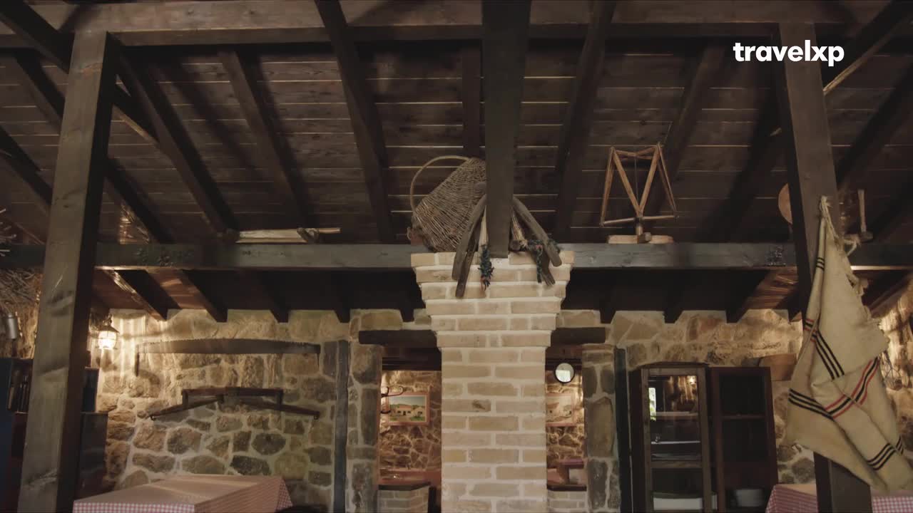 Under a dark wooden ceiling, a rustic interior of stone and brick is visible. Woven baskets and wooden tools rest atop a central brick pillar, while a checkered tablecloth covers a table below.
