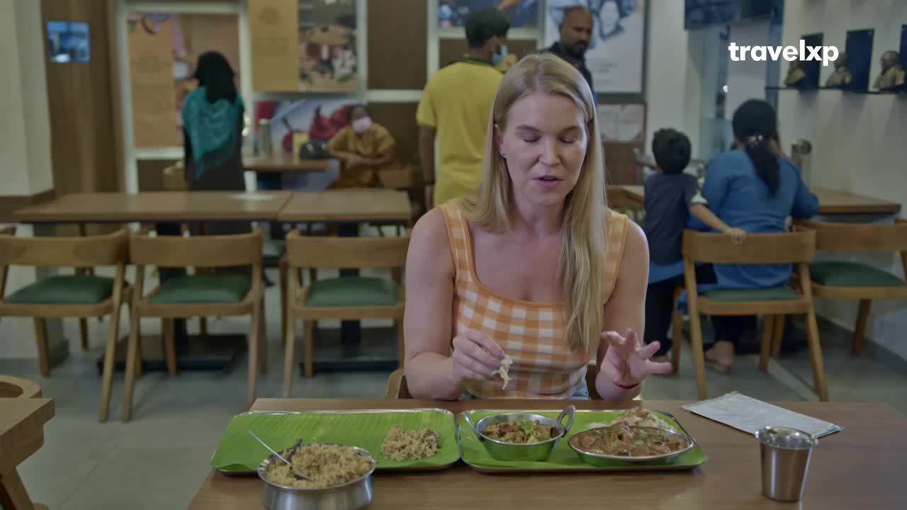 A woman in a checkered top sits at a table, holding a piece of food and gesturing with her other hand. Several dishes of food are laid out in front of her, and other diners are visible in the background of this restaurant in India.
