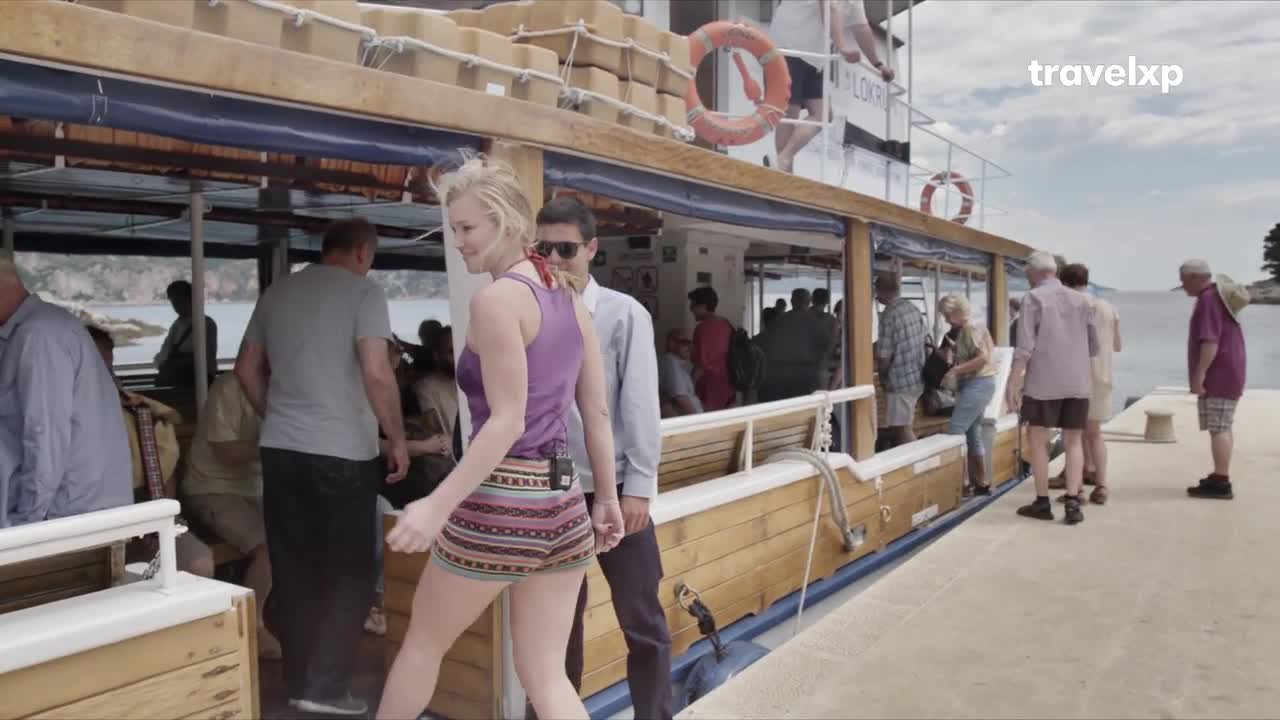 People are disembarking from a boat onto a concrete pier. A woman in a purple tank top and patterned shorts strides confidently toward the camera.
