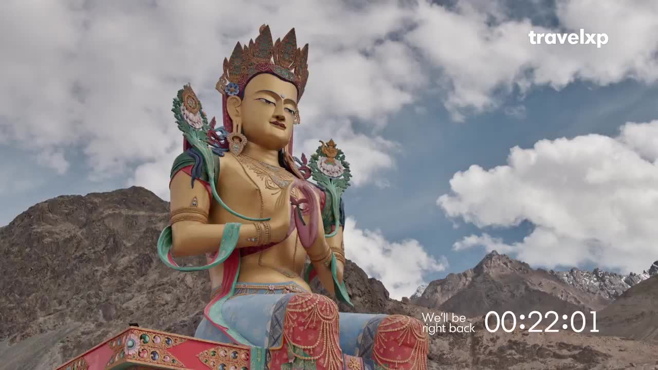 A large, golden statue of a seated Buddha dominates the frame, set against a backdrop of mountains and a cloudy sky. The Travelxp logo and a countdown timer signal a break in the broadcast, likely from India.
A large, golden statue of a seated Buddha dominates the frame, set against a backdrop of mountains and a cloudy sky. The Travelxp logo and a countdown timer signal a break in the broadcast, likely from India.