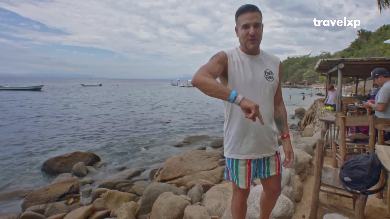 A man in a white tank top and striped shorts gestures towards the water, where small boats bob gently. Behind him, a beachside restaurant bustles with activity under a thatched roof.
A man in a white tank top and striped shorts gestures towards the water, where small boats bob gently. Behind him, a beachside restaurant bustles with activity under a thatched roof.