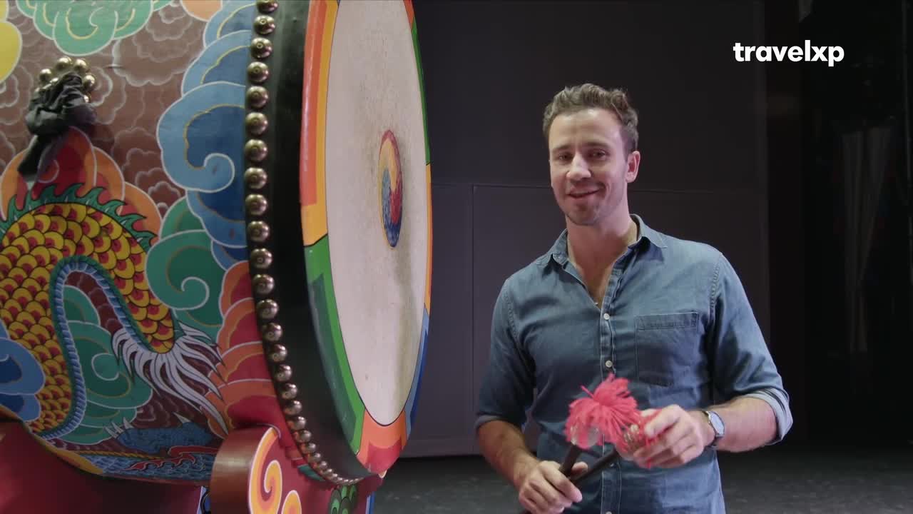 A man in a denim shirt stands beside a large, ornately painted drum. He holds a red, fluffy mallet, ready to play.
A man in a denim shirt stands beside a large, ornately painted drum. He holds a red, fluffy mallet, ready to play.