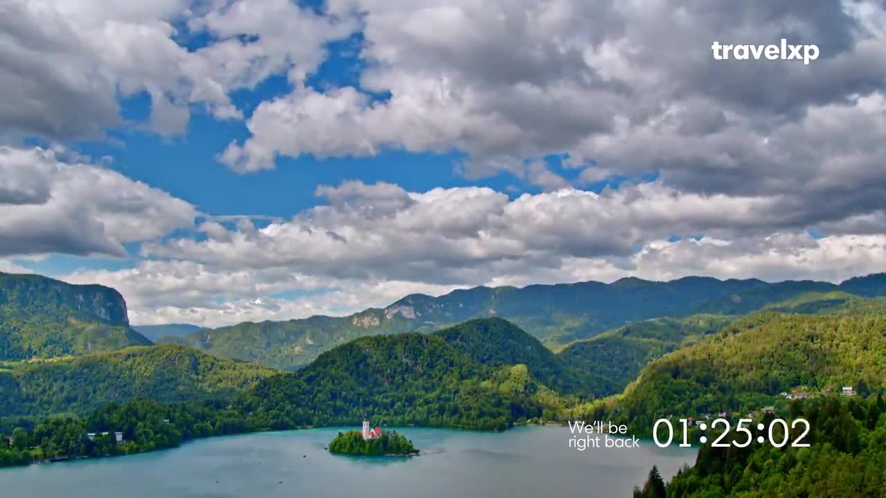 A vast lake reflects the sky, dotted with a tiny island church. Above, the clouds are thick, but patches of blue peek through, and the Travelxp logo is in the corner.
A vast lake reflects the sky, dotted with a tiny island church. Above, the clouds are thick, but patches of blue peek through, and the Travelxp logo is in the corner.