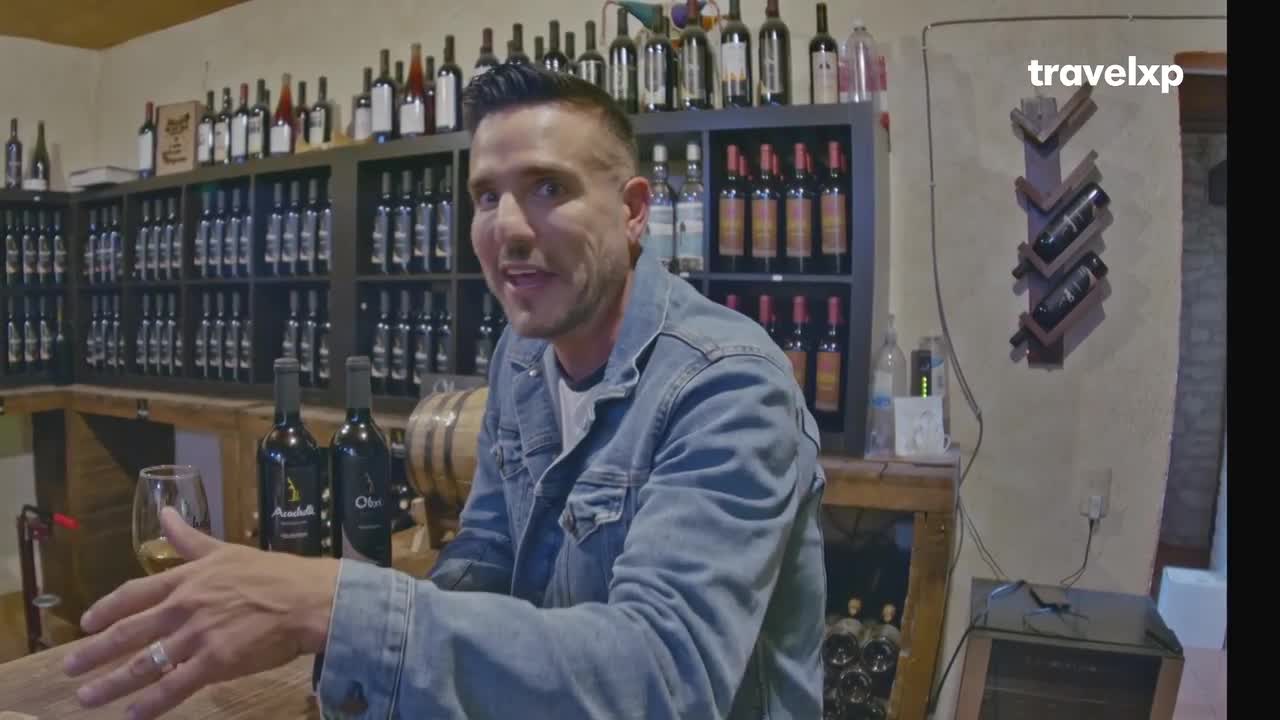 A man in a denim jacket gestures with a glass of wine, speaking towards the camera. Behind him, shelves are packed with bottles in a tasting room, the Travelxp logo visible in the corner.
A man in a denim jacket gestures with a glass of wine, speaking towards the camera. Behind him, shelves are packed with bottles in a tasting room, the Travelxp logo visible in the corner.