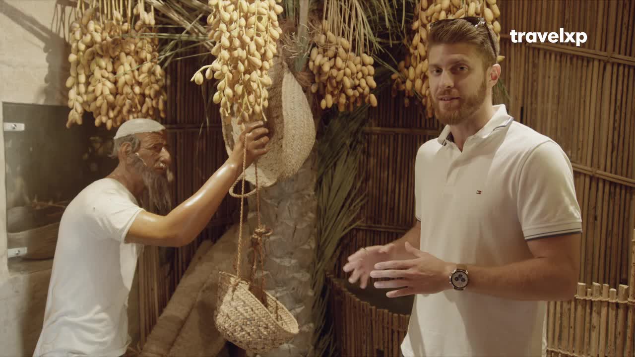 A man in a white polo shirt gestures towards a display of dates, while a statue of an older man with a long beard appears to be harvesting them. The Travelxp logo is visible in the corner.
A man in a white polo shirt gestures towards a display of dates, while a statue of an older man with a long beard appears to be harvesting them. The Travelxp logo is visible in the corner.