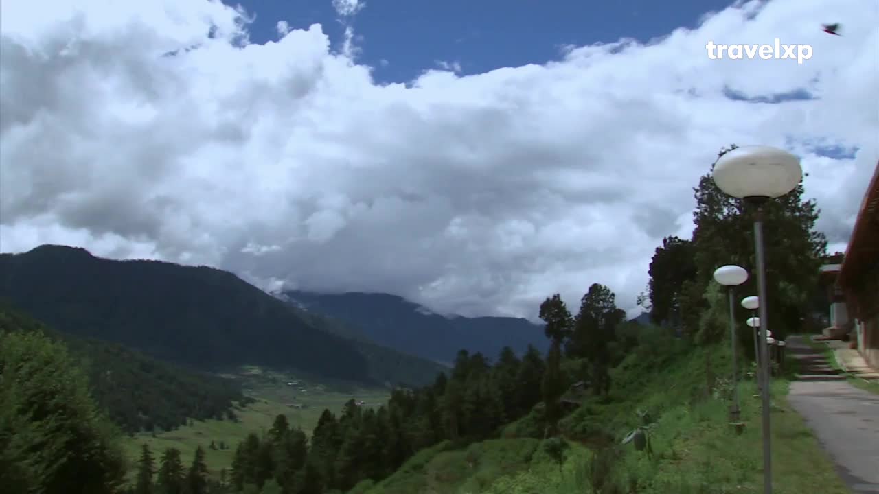 A vast valley unfolds between dark, tree-covered mountains under a sky filled with dramatic clouds. A pathway lined with modern lamps leads past a building on the right, hinting at a peaceful stop on a journey through India. A vast valley unfolds between dark, tree-covered mountains under a sky filled with dramatic clouds. A pathway lined with modern lamps leads past a building on the right, hinting at a peaceful stop on a journey through India.