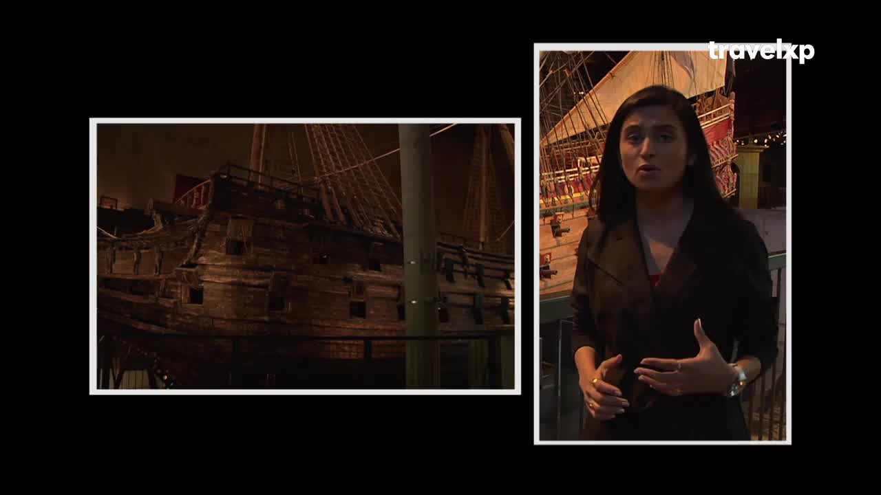 A woman stands before a historic ship, its wooden hull dark and imposing. She gestures as she speaks, a Travelxp logo visible in the background. A woman stands before a historic ship, its wooden hull dark and imposing. She gestures as she speaks, a Travelxp logo visible in the background.