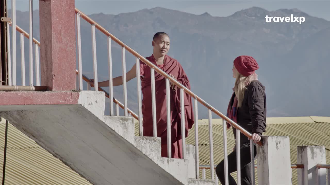 A monk in maroon robes stands on a staircase, gesturing as he speaks to a woman in a red hat. The backdrop of the Himalayas hints at a journey through India, perhaps captured for Travelxp. A monk in maroon robes stands on a staircase, gesturing as he speaks to a woman in a red hat. The backdrop of the Himalayas hints at a journey through India, perhaps captured for Travelxp.
