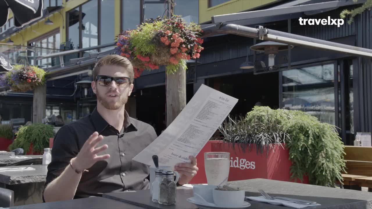 A man in sunglasses gestures with a menu while speaking at an outdoor restaurant. A Travelxp logo appears in the upper right corner.