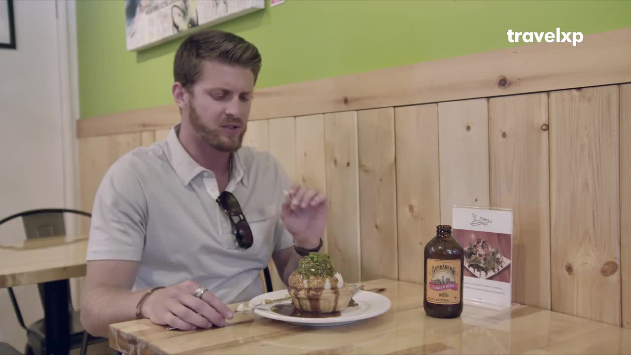 A man gestures with a fork over a dessert, a brown bottle and a menu resting nearby. This scene unfolds in a casual eatery, perhaps during a Travelxp segment exploring local flavors.