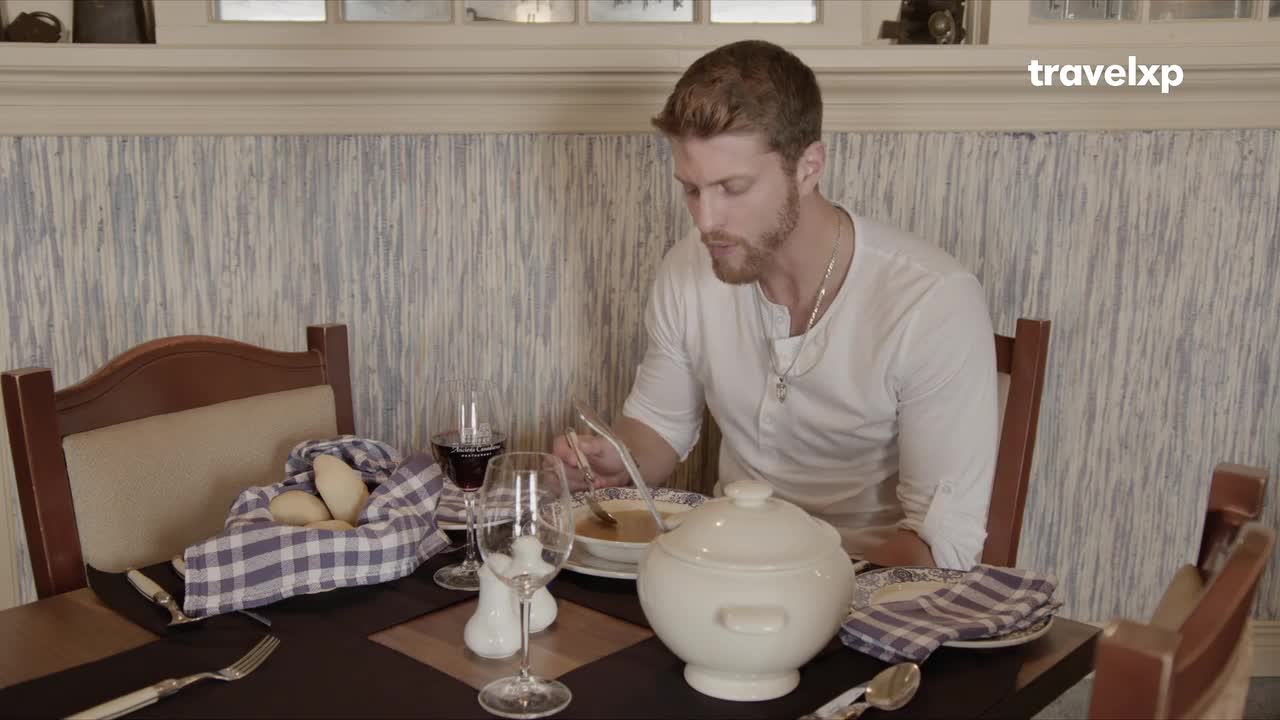 A man with a beard is eating soup at a table set for a meal. The Travelxp logo is visible in the corner of the frame.