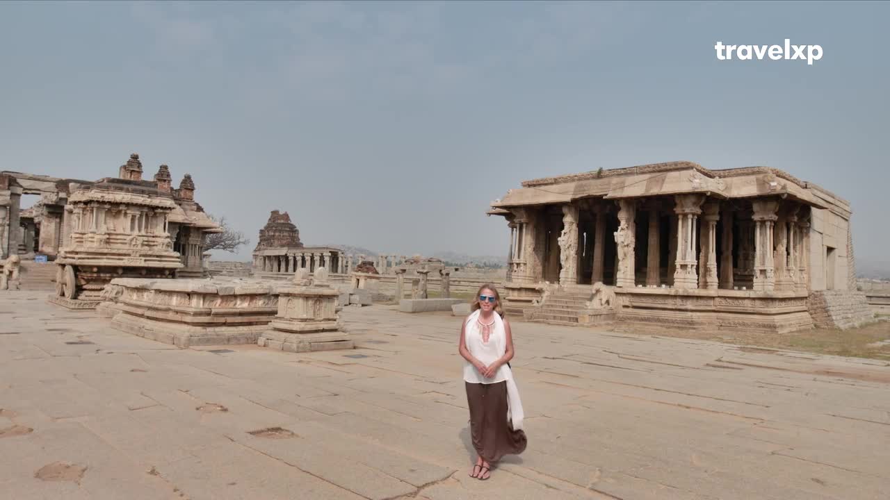A woman stands in the vast courtyard of an ancient Indian temple complex. Stone structures, including a chariot and pillared halls, stretch across the dusty landscape under a pale sky.