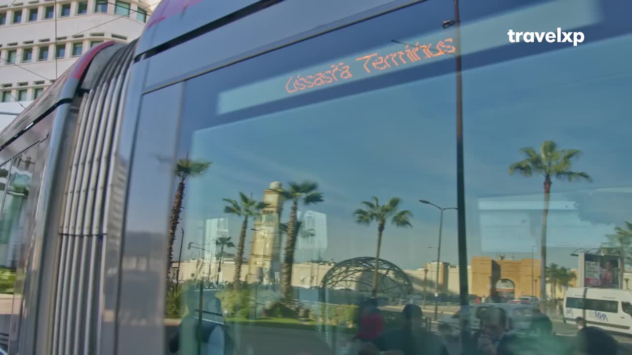 A modern tram glides past, its windows reflecting a bright blue sky dotted with palm trees and a distant archway. The tram's destination, "Casasra Terminus," glows in orange light on its digital display.
