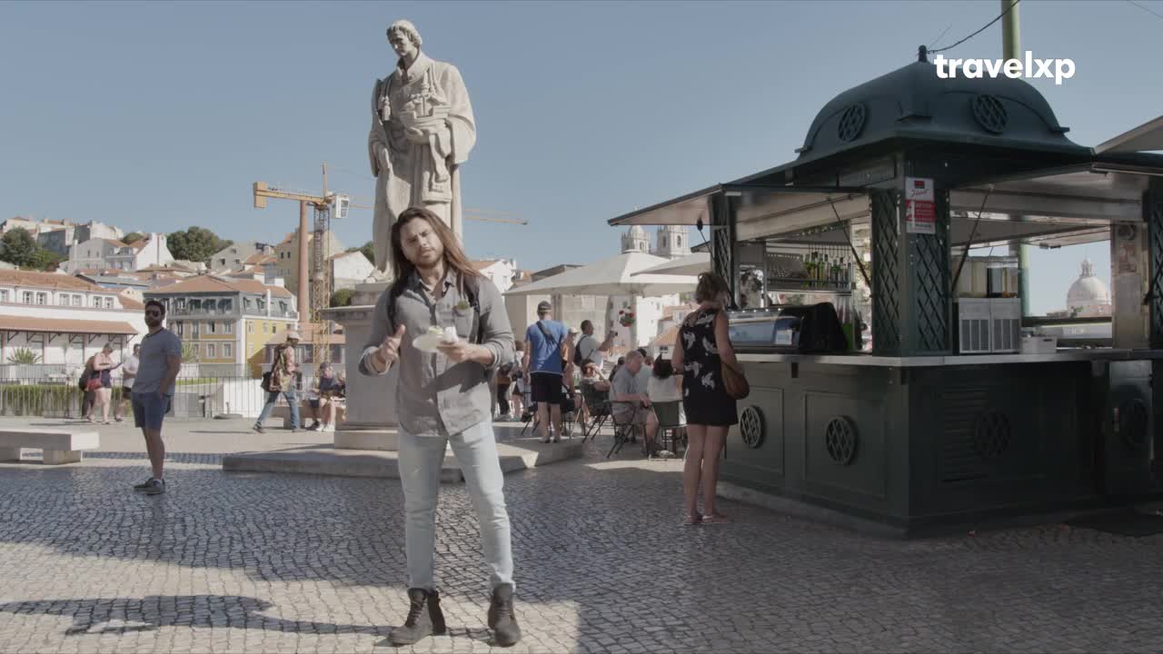 A man with long hair holds a small cup and gives a thumbs-up, standing near a statue. Behind him, people mill about a sunny plaza with a kiosk serving drinks.