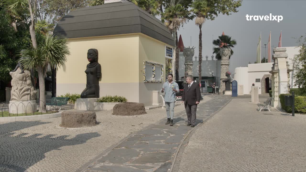 Two men walk along a stone path, one in a suit and the other in a light blue shirt. They are surrounded by unusual sculptures and palm trees under a bright sky.