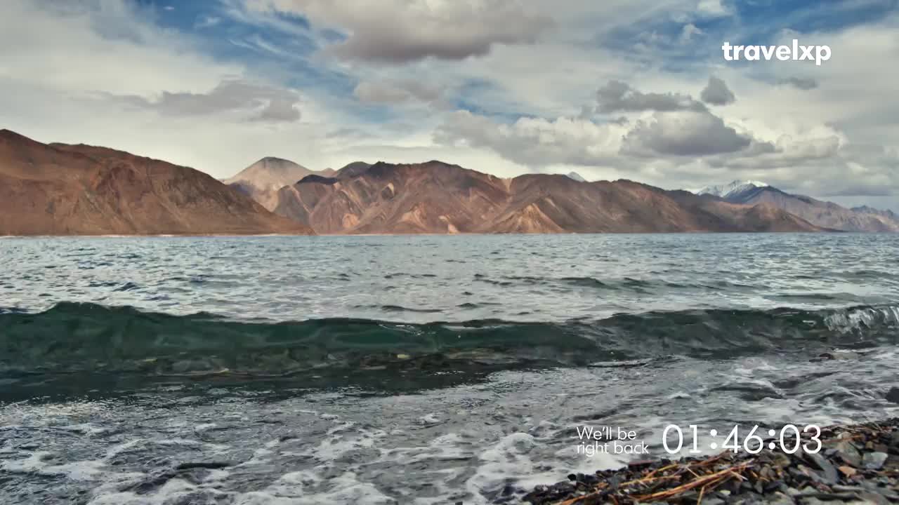 Waves lap gently against the rocky shore of a vast, blue lake. Jagged, brown mountains rise in the distance under a dramatic sky.