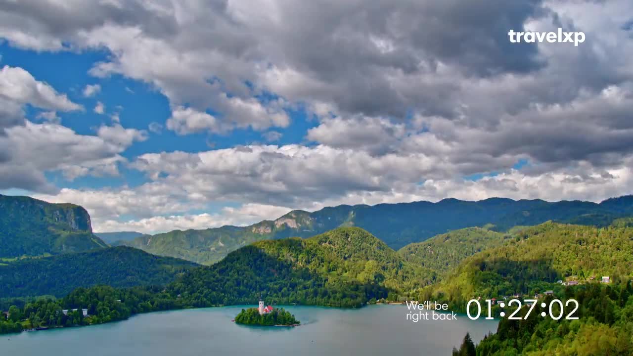 A small island church sits in a serene lake, surrounded by rolling green hills under a dramatic sky. The Travelxp logo is visible in the upper right corner.