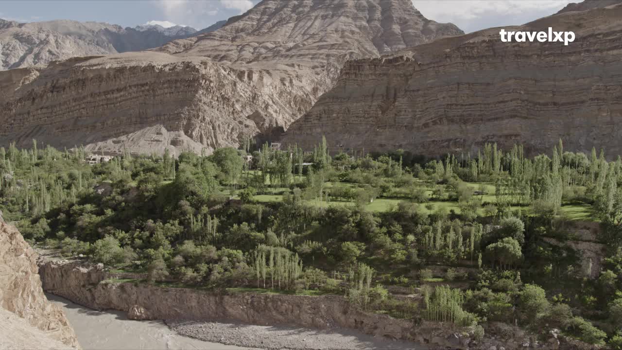 A wide, muddy river flows through a valley, its banks lined with steep, rocky slopes. Lush green trees and fields dot the landscape below the towering, layered mountains.