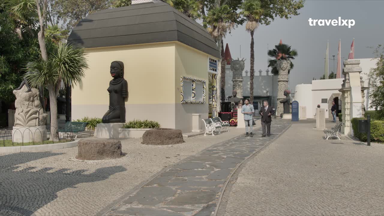 Two men walk along a stone path past a kneeling statue. A small train sits behind them, waiting for passengers.