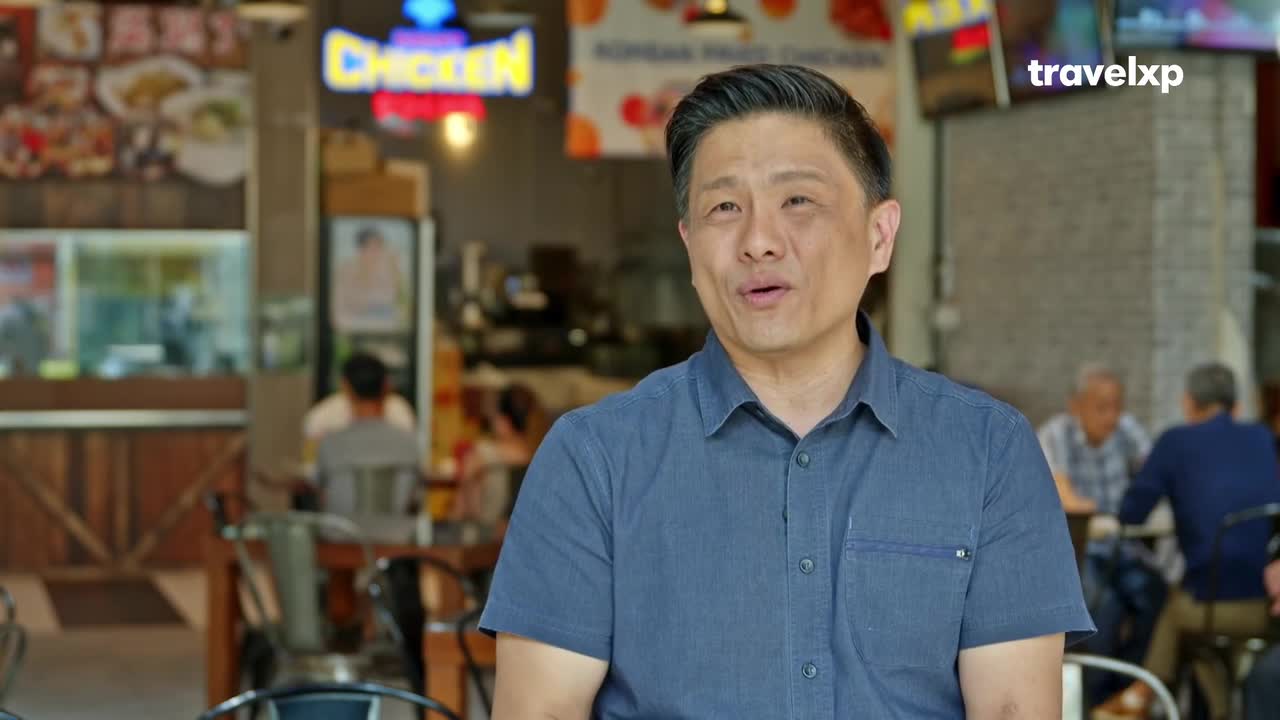 A man in a blue shirt stands in front of a bustling food court. Behind him, a neon sign for "CHICKEN" glows above a busy counter. People are seated at tables, and the general atmosphere suggests a lively dining experience.