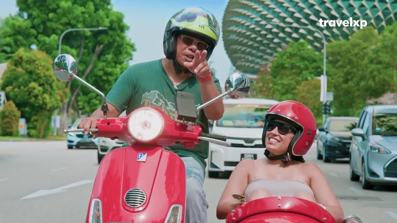 A man points forward while riding a red scooter, his companion smiling beside him. Cars pass by on a sunny day, with a distinctive architectural structure visible in the background.