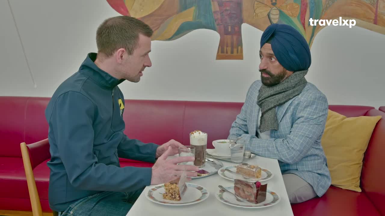 Two men sit at a table, sharing coffee and cake. The man in the blue jacket gestures as he speaks, while the man in the turban listens intently.