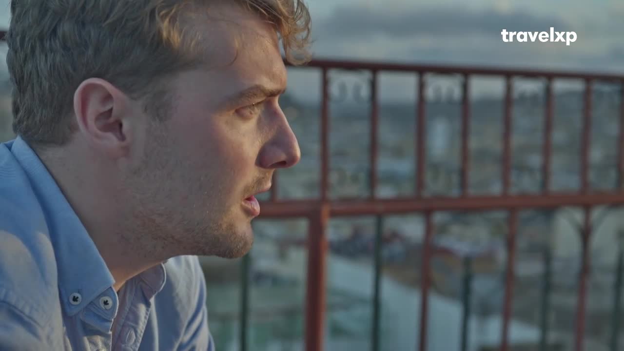 A man with light hair, wearing a blue shirt, looks out over a city skyline. The Travelxp logo is visible in the upper right corner.