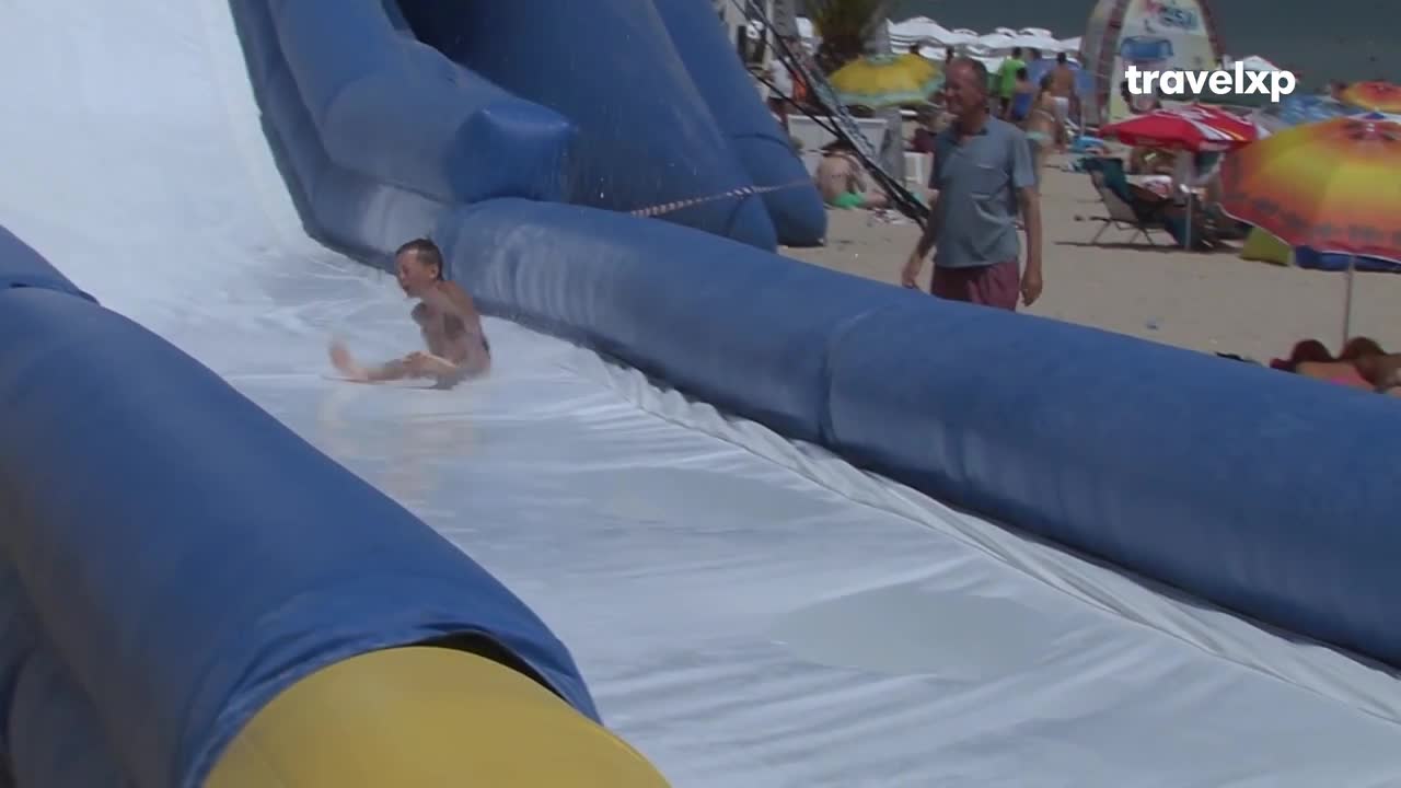 A young boy slides down a water slide at a beach resort, his legs kicking up spray. Further up the slide, a man watches the descent, a relaxed smile on his face as the Travelxp logo appears over the scene.