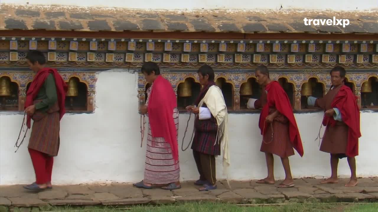 A line of men in traditional dress walks along a white wall adorned with colorful patterns and small bells. Each man carries a string of prayer beads, their steps steady on the stone path.