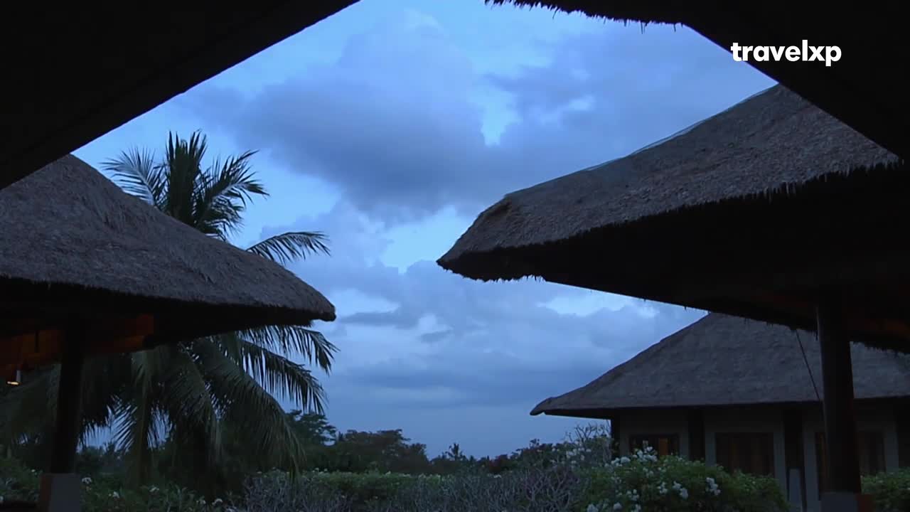 The sky darkens over thatched roofs and palm trees. A Travelxp logo appears in the upper right corner.