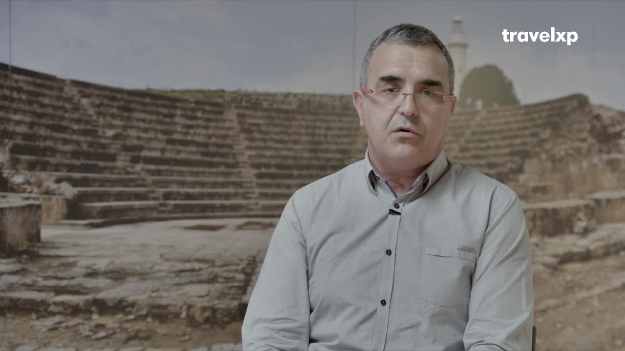 A man in a light blue shirt speaks directly to the camera, his gaze steady. Behind him, the weathered stone tiers of an ancient amphitheater rise, a silent testament to history.